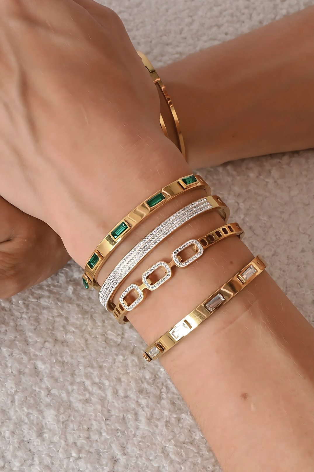 Close-up of a wrist wearing multiple gold, silver, and green gemstone bracelets on a neutral background.
