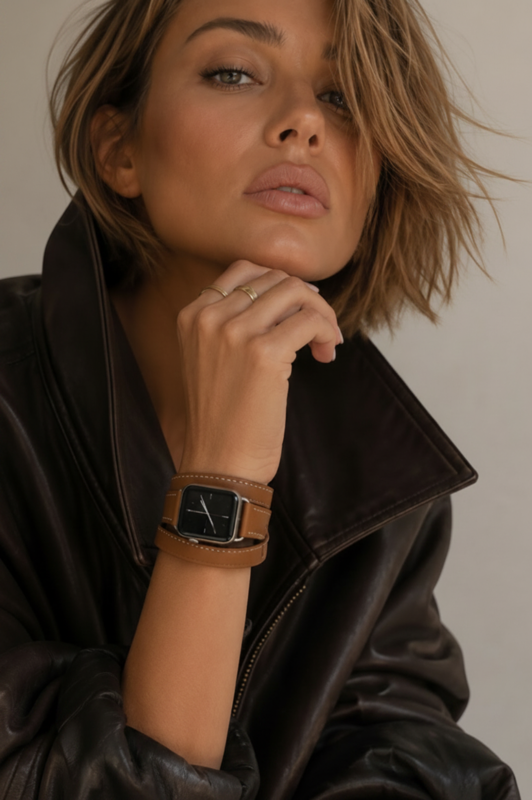 A close-up lifestyle shot of a woman wearing a brown leather watch with a square black dial and a matching brown leather strap, styled with a dark brown leather jacket.