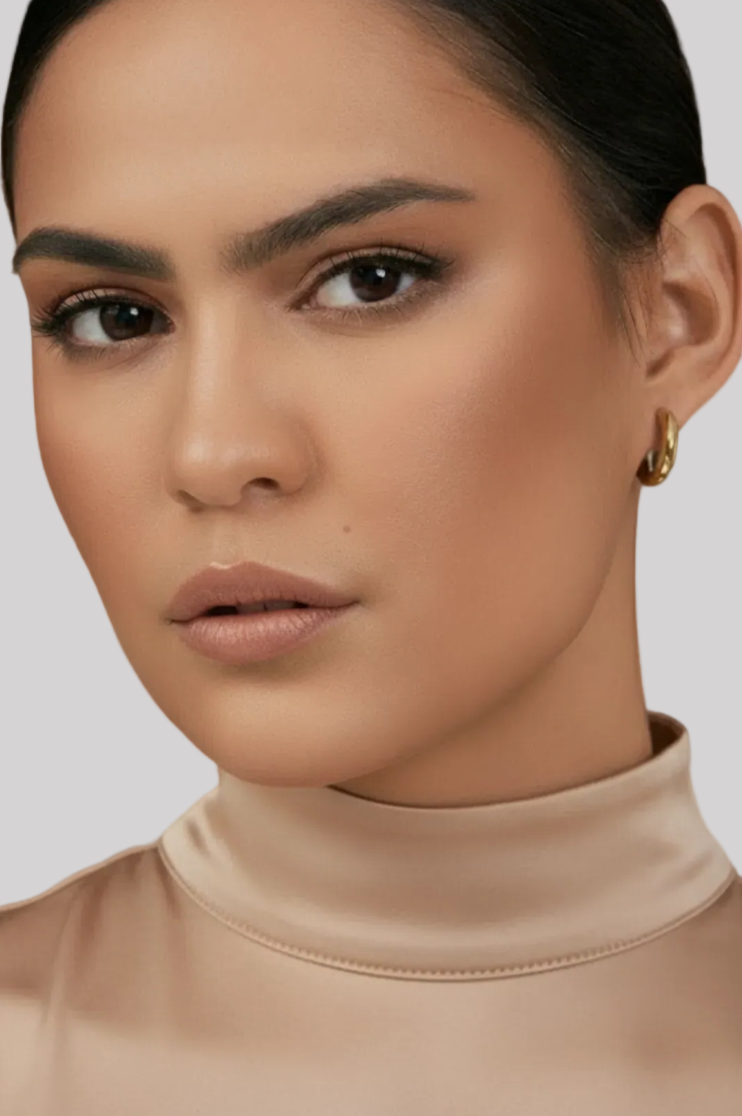 Close-up of a woman wearing a beige turtleneck and gold earrings against a white background